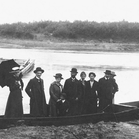 Powiększ obraz: Archiwalne zdjęcie przedstawiające elegancko ubranych ludzi stojących w łodzi, za nimi znajduje się tratwa-prom